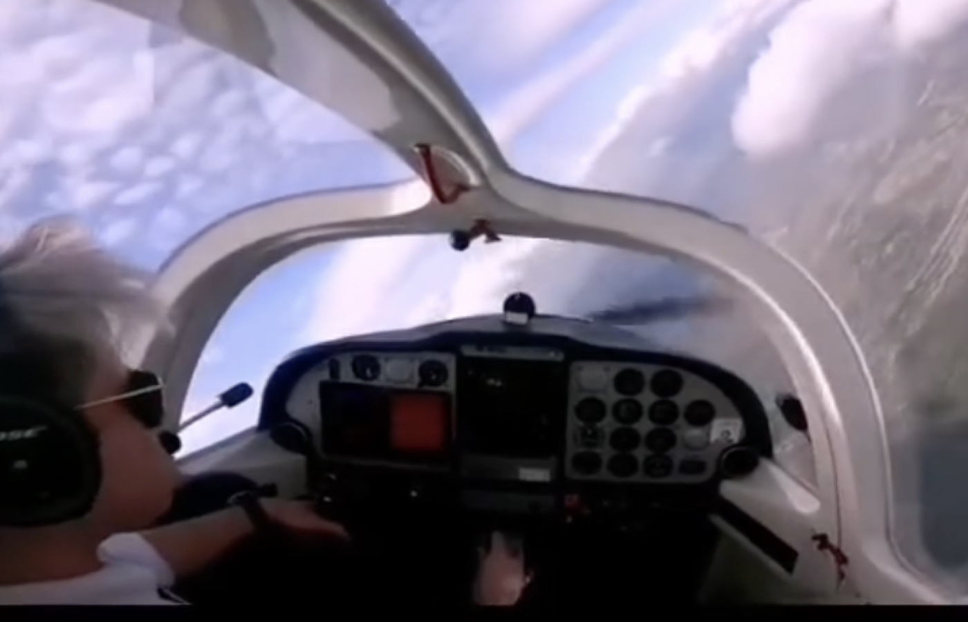 Jean flying a small aircraft, sunglasses on, clouds filling the canopy