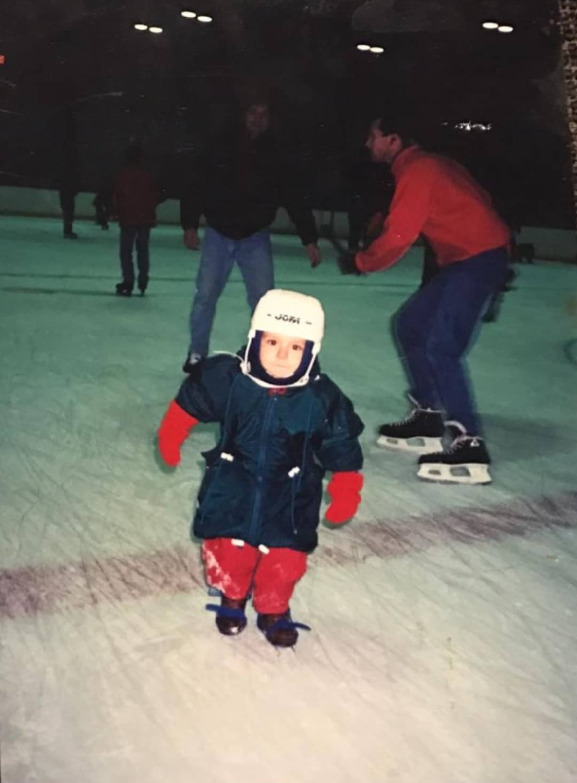 Jean as a toddler on a skating rink, bundled in a snowsuit and helmet, taking his first steps on the ice