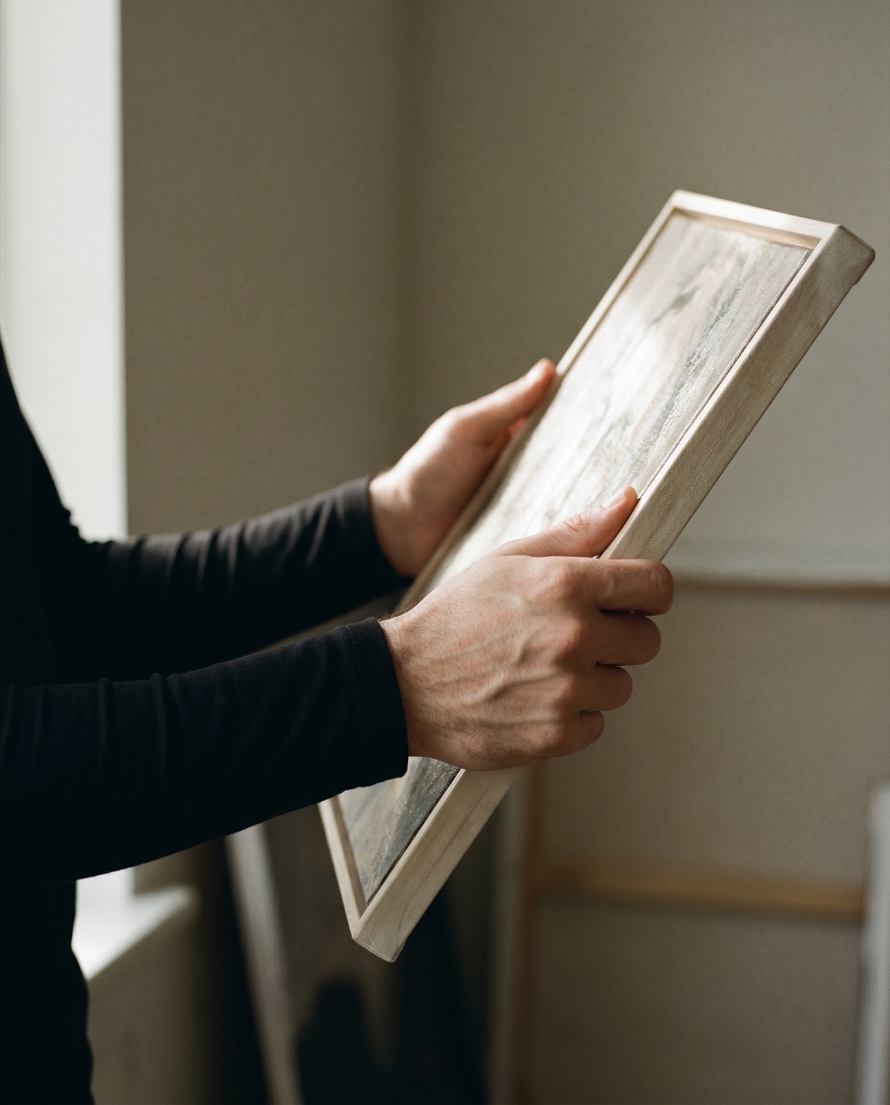 Hands holding a small canvas up to the window light to examine it
